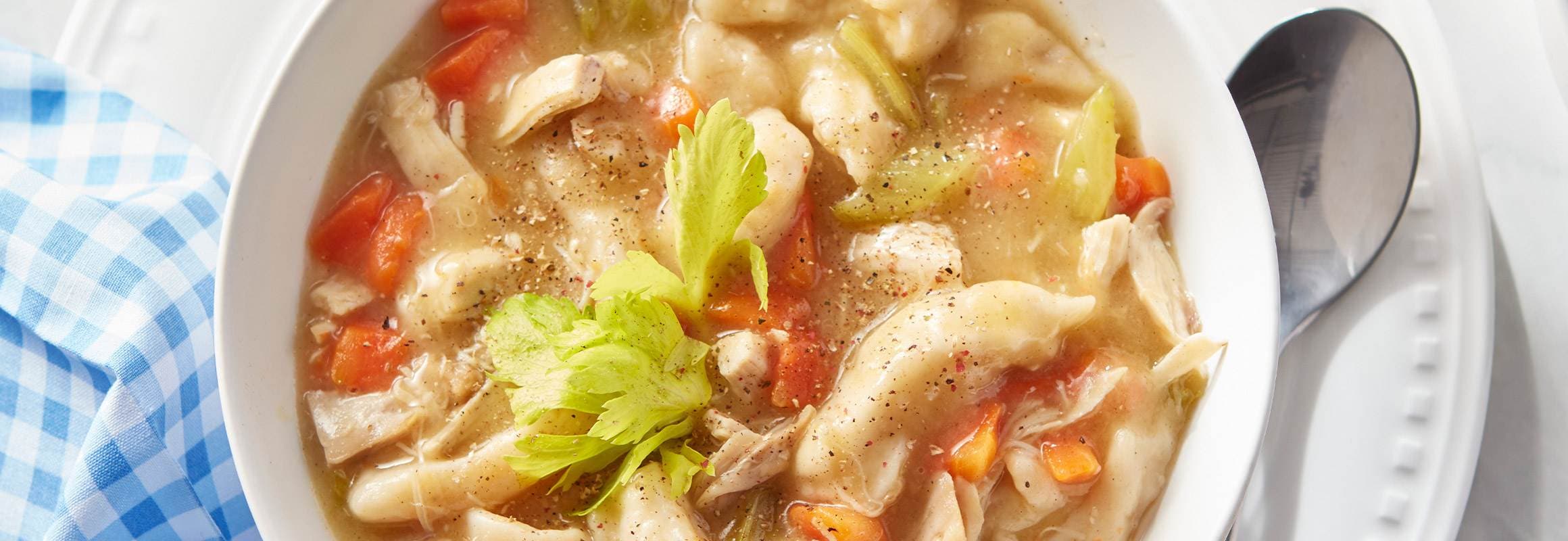 A bowl of chicken and dumplings soup with carrots and celery, garnished with a celery sprig and black pepper.