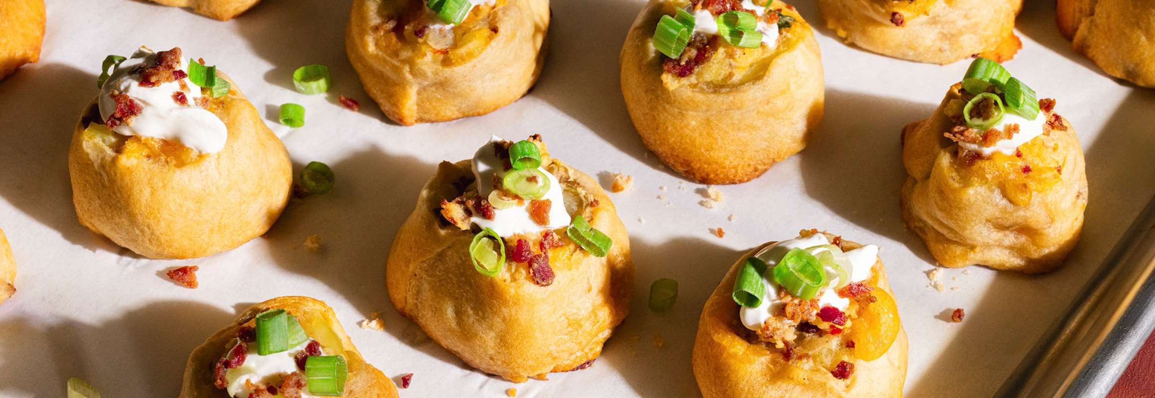White serving plate with pinwheels filled with potatoes, cheese, and bacon topped with green onion and sour cream. In the background there’s a small sauce dish filled with sour cream.