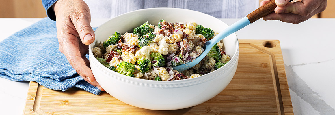 A person holding a white bowl filled with Sunny Broccoli Pasta Salad on a wooden cutting board. The salad includes broccoli florets, radiatore pasta, bacon bits, and cauliflower, with a creamy dressing. A blue cloth napkin is visible in the background